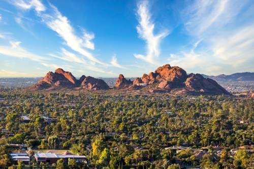 A panoramic view of red rock formations surrounded by lush greenery and a blue sky with wispy clouds.