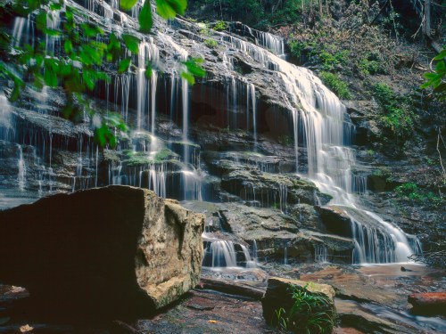 A serene waterfall cascading over rocky cliffs, surrounded by lush greenery and a large boulder in the foreground.