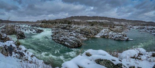 A panoramic view of a snowy river landscape with rocky outcrops and swirling turquoise waters under a cloudy sky.