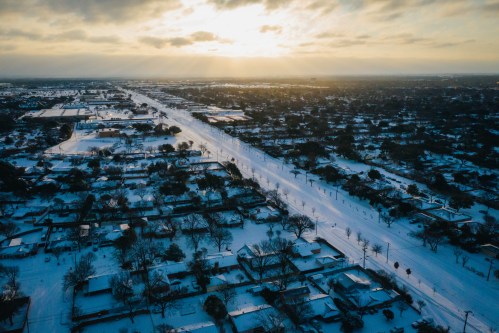 Aerial view of a snowy landscape with houses and a road, under a cloudy sky at sunset.