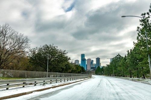 A snowy road leads towards a city skyline under a cloudy sky, with trees lining the sides.
