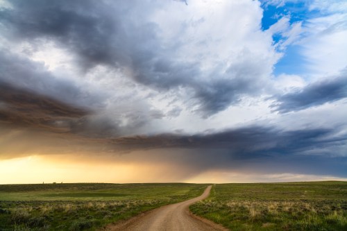 A winding dirt road stretches through a grassy landscape under a dramatic sky filled with clouds and sunlight.