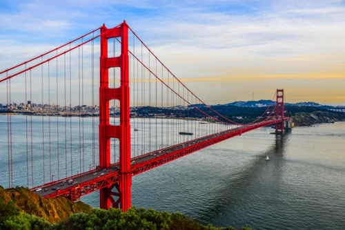 Golden Gate Bridge spans a bay, with a vibrant red color against a backdrop of water and hills under a blue sky.