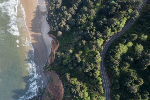 Aerial view of a coastline with waves, sandy beach, and a winding road surrounded by lush greenery.