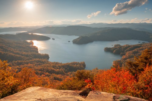A scenic view of a lake surrounded by autumn foliage and mountains under a bright sky with the sun setting.