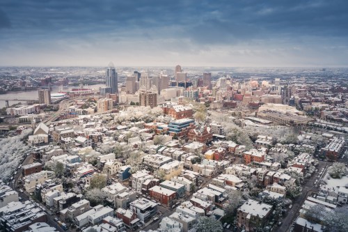 Aerial view of a snowy cityscape with buildings and trees, showcasing a skyline under a cloudy sky.