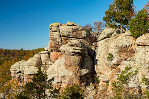 Rocky cliffside with layered stone formations and sparse trees against a clear blue sky.