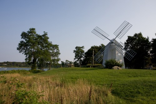 A historic windmill stands on a grassy area near a calm body of water, surrounded by trees under a clear blue sky.