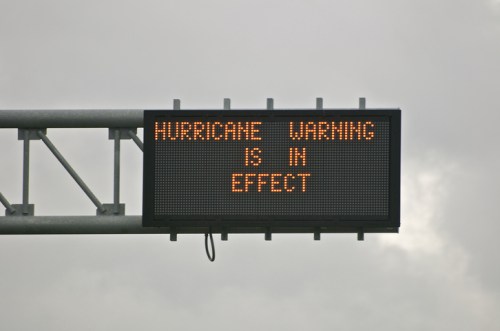 Digital road sign displaying "HURRICANE WARNING IS IN EFFECT" against a cloudy sky.