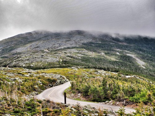A winding road curves through a mountainous landscape, with clouds hovering over the rocky peak and lush greenery below.