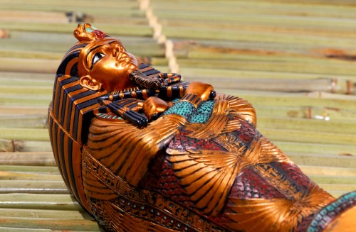 A detailed, ornate golden sarcophagus resembling an ancient Egyptian pharaoh, resting on a bamboo surface.