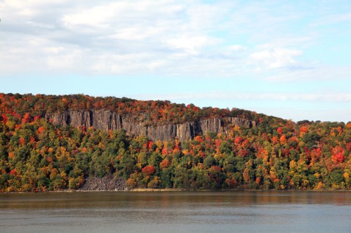 A scenic view of a river with a colorful autumn forest and rocky cliffs in the background under a partly cloudy sky.