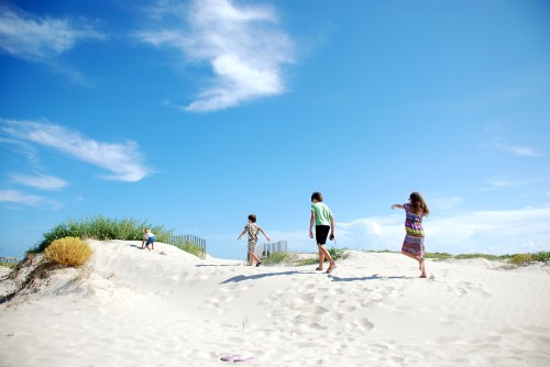 Four children play on sandy dunes under a bright blue sky with scattered clouds.