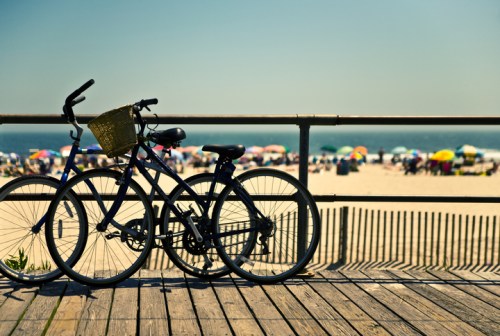 Two bicycles parked on a wooden boardwalk, with a beach and colorful umbrellas in the background.