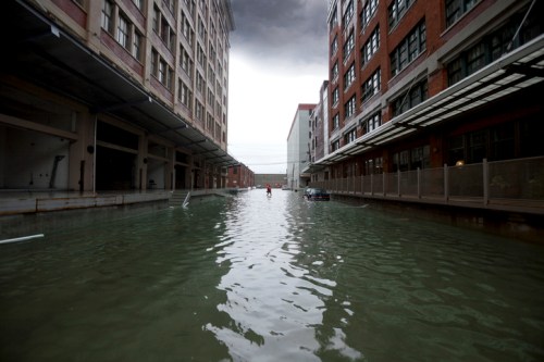 A flooded urban street with water covering the ground, surrounded by tall buildings under a cloudy sky.