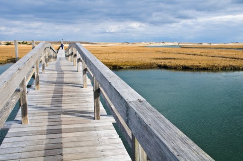 A wooden pier extends over calm water, leading into a grassy landscape under a cloudy sky.