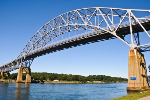 A large steel arch bridge spans a calm river, surrounded by green trees and a clear blue sky.