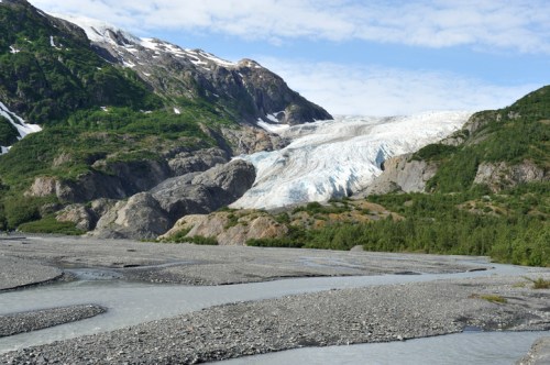 A glacier flows down a mountain, surrounded by green hills and a riverbed of rocks under a blue sky.