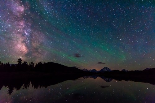 A starry night sky with the Milky Way, reflecting on a calm lake surrounded by mountains and trees.
