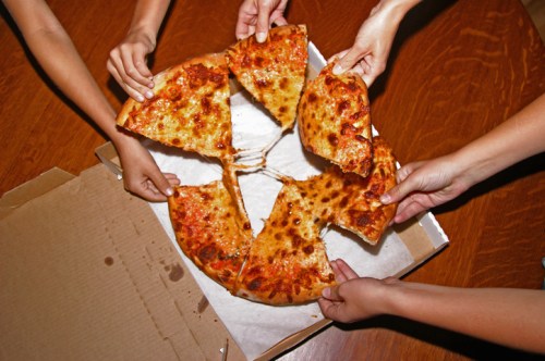 Several hands reaching for slices of a cheese pizza in a cardboard box on a wooden table.