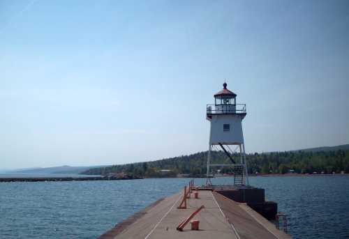 A white lighthouse stands on a pier, surrounded by calm water and green hills under a clear blue sky.