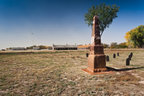 A historic site featuring a stone monument, grassy field, and old buildings under a clear blue sky.