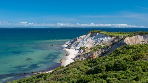 A scenic coastal view featuring white cliffs, a sandy beach, and lush greenery under a clear blue sky.