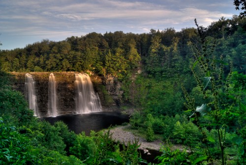 A scenic waterfall cascades into a serene pool, surrounded by lush green trees and a clear blue sky.