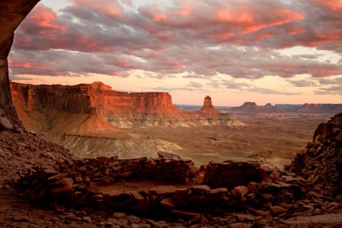 Sunset over a vast desert landscape with rock formations and dramatic clouds, viewed from a cave.