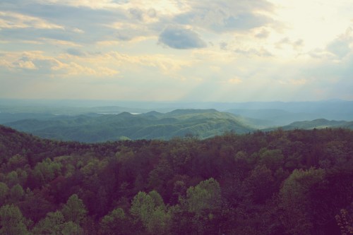 A panoramic view of rolling green mountains under a cloudy sky, with soft sunlight breaking through the clouds.
