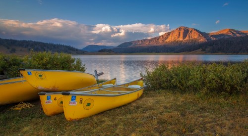 Three yellow canoes are docked by a serene lake, surrounded by lush greenery and mountains under a colorful sky.