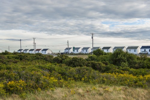 A row of white houses along a road, surrounded by green shrubs and under a cloudy sky.