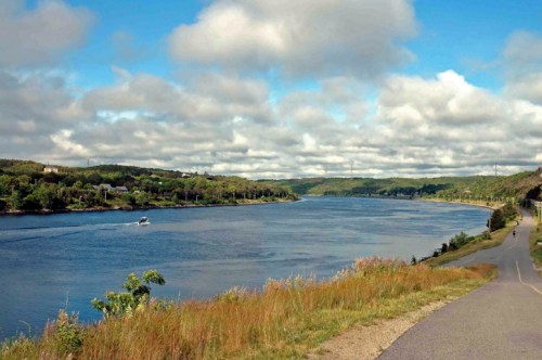 A serene river scene with a boat on the water, surrounded by green hills and a cloudy blue sky.