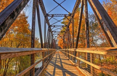 A wooden bridge with metal supports spans a river, surrounded by vibrant autumn foliage under a clear blue sky.