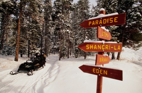 A snowmobile parked near a wooden signpost with directional arrows to "Paradise," "Shangri-La," "Nirvana," and "Eden."