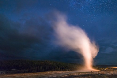 A geyser erupts at night, sending a plume of steam and water into the starry sky, surrounded by dark trees.