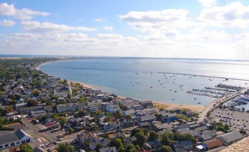 Aerial view of a coastal town with boats in the water and a sandy beach under a blue sky with scattered clouds.