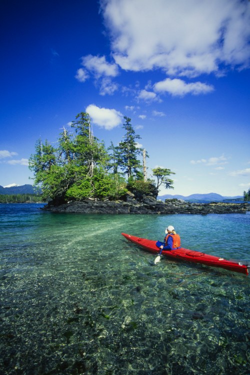 A person in a red kayak paddles in clear water near a small island with trees under a blue sky.