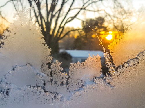 Frosty window with intricate ice patterns, framing a sunrise over a winter landscape.