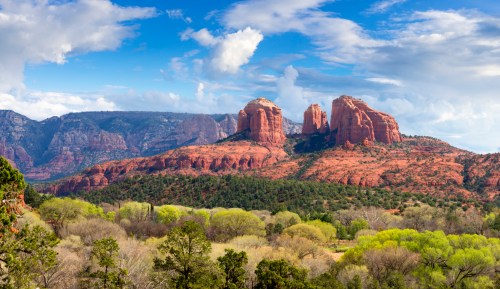 A scenic view of red rock formations under a blue sky with clouds, surrounded by green trees and mountains in the background.