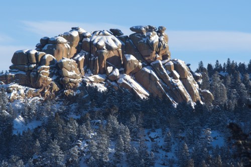 Snow-covered rocky outcrop surrounded by evergreen trees under a clear blue sky.