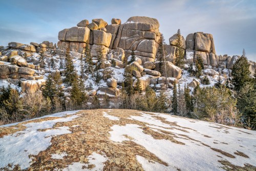 Rocky outcrop surrounded by snow and evergreen trees under a clear sky.