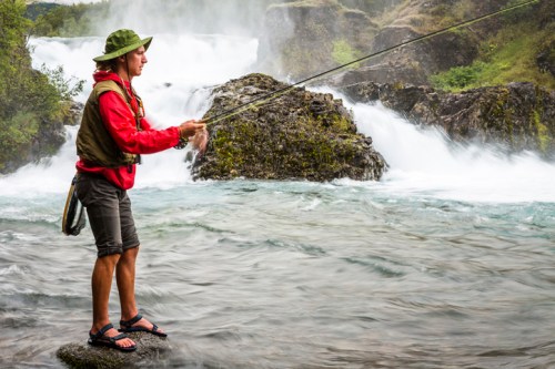 A person in a red jacket and hat stands on a rock, fishing by a rushing waterfall in a scenic outdoor setting.