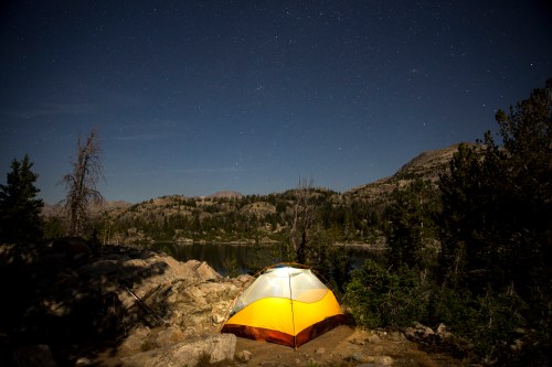 A brightly colored tent sits by a serene lake under a starry night sky, surrounded by mountains and trees.