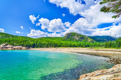A scenic beach with clear turquoise water, surrounded by lush green hills and a bright blue sky with fluffy clouds.