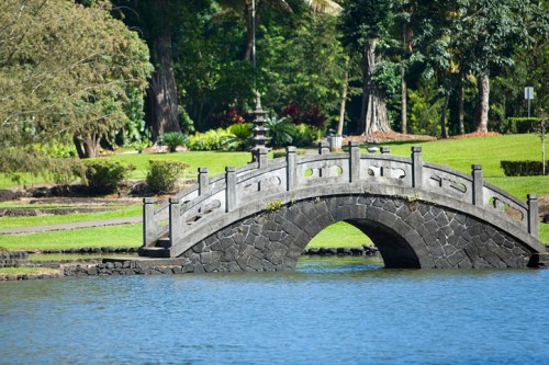 A stone bridge arches over a calm lake, surrounded by lush greenery and trees in a serene park setting.