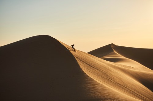 A silhouette of a person climbing a sand dune at sunset, with soft, flowing sand formations in the background.