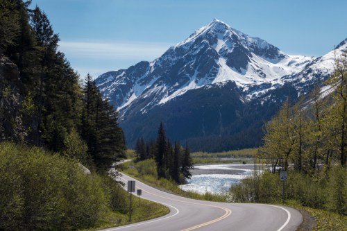 A winding road leads through lush greenery, with a snow-capped mountain and a river in the background under a clear blue sky.