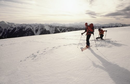Two skiers navigate a snowy mountain landscape under a cloudy sky, with rugged peaks in the background.