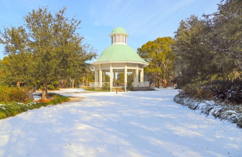 A white gazebo with a green roof surrounded by snow-covered ground and trees in a serene park setting.
